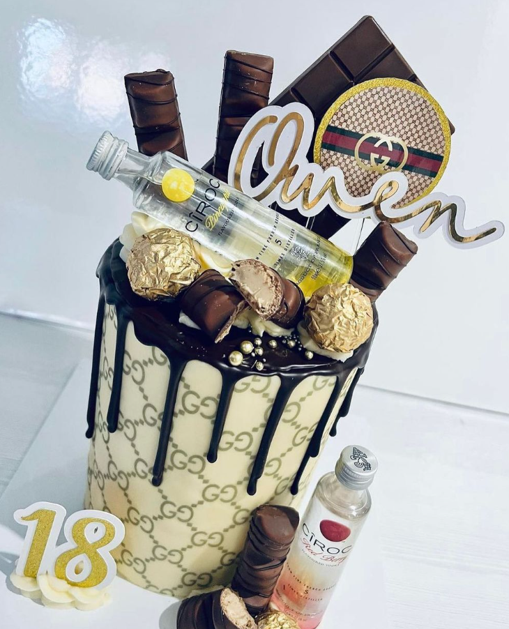 gucci printed cake wrap around a tall iced cake with chocolate drip. Decorations include vodka, chocolate bars and non edible deals with a name and age.