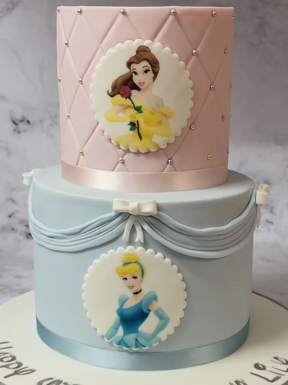 2 Tier Disney princess birthday cake, the top tier is iced in a pink quilted effect with an edible image of Belle. The bottom tier is blue with icing draped around the top and ribbon around the bottom. The bottom tier has an edible image of Cinderella.