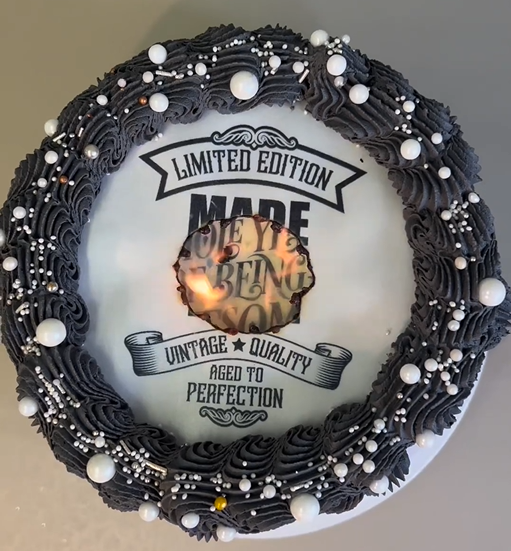 a burn away cake in the process of burning showing glimmer of the reveal image. The cake is decorated with black piped buttercream icing with while pearls sprinkled over it