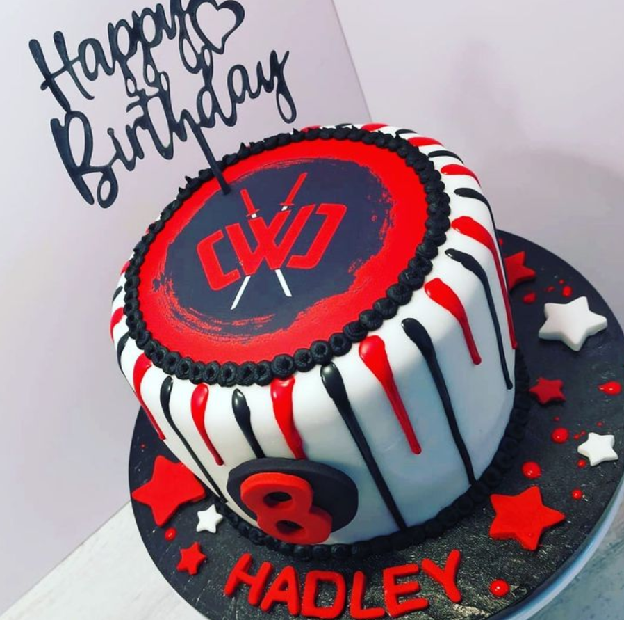 Black and red cake with a drip effect. White and red stars are scattered on the cake board. An edible circle icing sheet, printed with a black and red logo is on the top of the cake, surrounded by black piping.