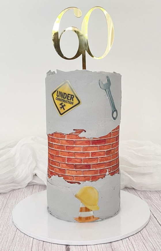 Builders 60th birthday cake, topped with an acrylic number 60. Edible images on the side of the cake consist of an 'under construction' sticker, a spanner, a hard hat and cone. Arounf the middle of the cake in an edible printed stone wall which surrounds the cake.