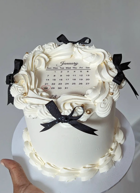 tall ivory cake iced with ivory buttercream and black ribbon bows. In the centre is an edible calender print with the date encased in a heart and a personalised message