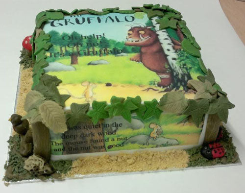 Square cake in a Gruffalo theme. The edges are adorned with green icing foliage and the cake board has grass and sand effects. The top and the sides of the cakes show edible images of the Gruffalo and phrases with different characters