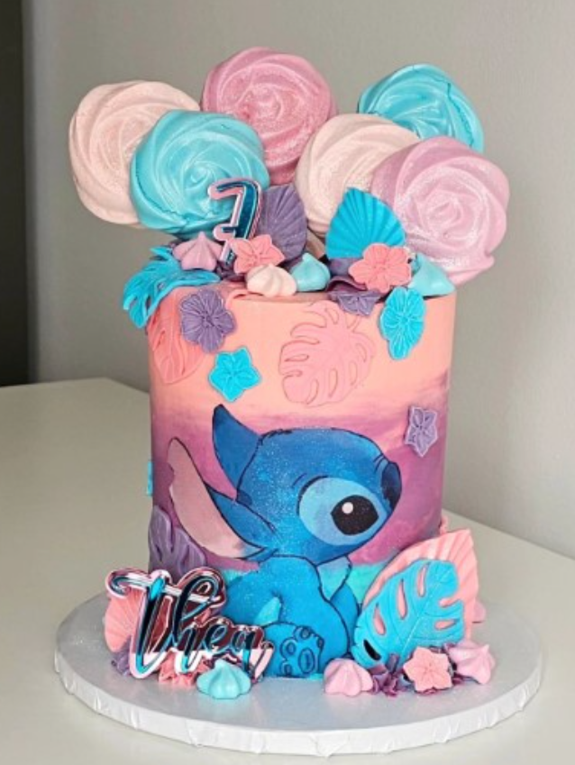 Disney Stitch cake in pink, peach and blue. Edible roses in the same colours are sat on the top of the cake with foliage draping down the side. On the front of the cake is an icing image of stitch.
