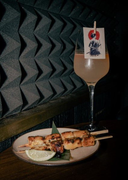 chicken skewers on a plate with a tall glass of rose champagne, attached to the glass with a wooden peg is an edible rectangle wafer card with a custom printed image.