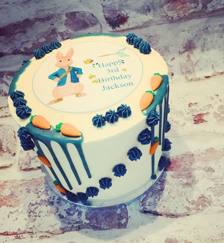 Peter Rabbit inspired cake with icing shaped carrots and blue butter cream piping sporadically placed on the top and sides. An edible printed circle is in the center of the cake with Peter rabbit and a birthday message.