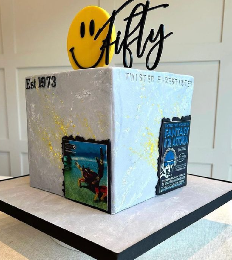 tall square cake with a grey marbled effect icing, on each side there is text made from icing with says Est 1973 with an edible photo or logo. The top of the cake has an acrylic topper with a yellow emoji smiley face and the word 'Fifty'.